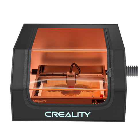 A laser engraver inside a black enclosure with a transparent window showing the engraving process for real-time observation. The protective cover has the brand name "CrealityFalcon" displayed on the bottom front panel, and a cable is visible on the right side.