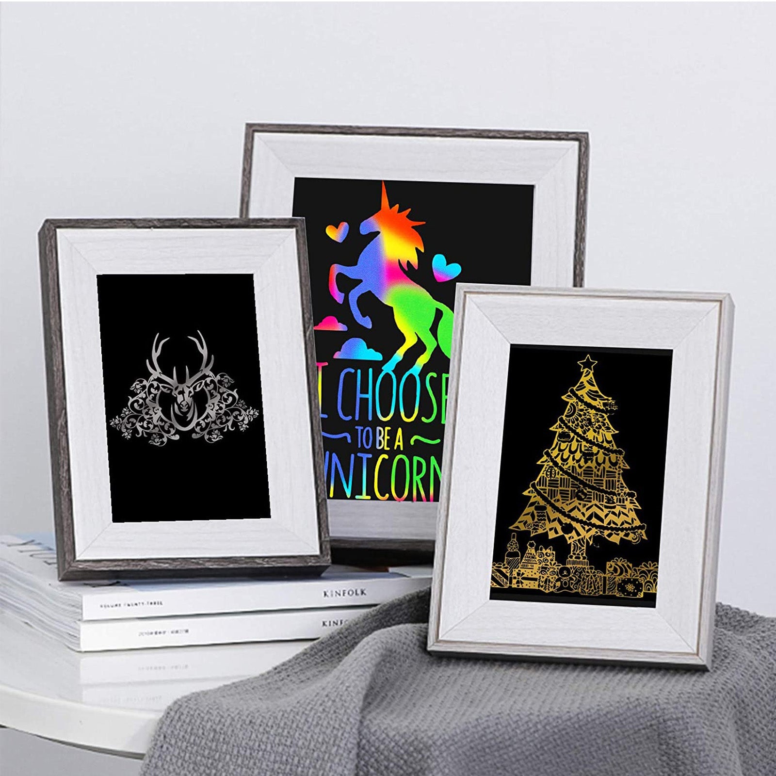 Three framed prints sit on a white table. The left frame features an intricate deer head sketch. The middle frame shows a colorful unicorn with the text "I choose to be a unicorn." The right frame displays a gold Christmas tree design, ideal for laser engraving ideas using CrealityFalcon's A4 Gold & Silver Scratch Paper for Laser Engraving - 40 PCS. A gray blanket is draped partly over the table.