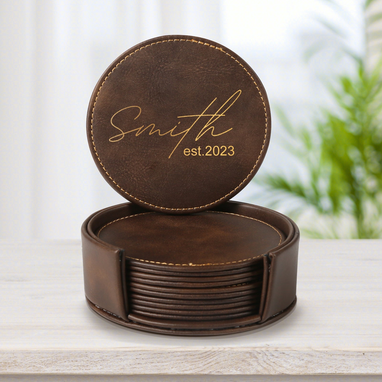 A hand holds a green mug with the text "Lucky" printed on it. The mug is placed near a set of CrealityFalcon Personalised Brown Leather Coasters with Holder for Falcon Home Laser Engraving - 6 PCS, one of which features intricate woodworking laser engraving and is out of the holder on a wooden table. A potted plant with green leaves is in the background.