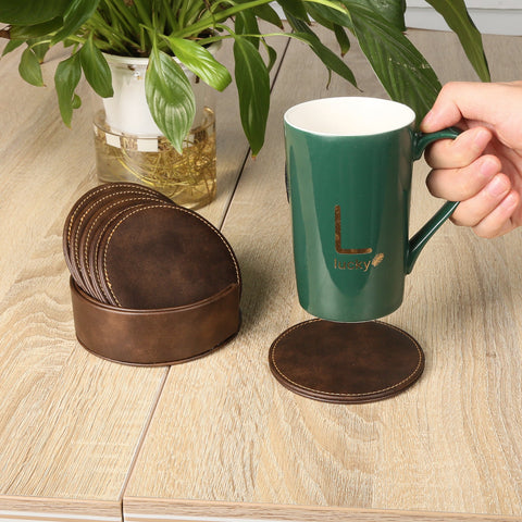 A hand holds a green mug with the text "Lucky" printed on it. The mug is placed near a set of CrealityFalcon Personalised Brown Leather Coasters with Holder for Falcon Home Laser Engraving - 6 PCS, one of which features intricate woodworking laser engraving and is out of the holder on a wooden table. A potted plant with green leaves is in the background.